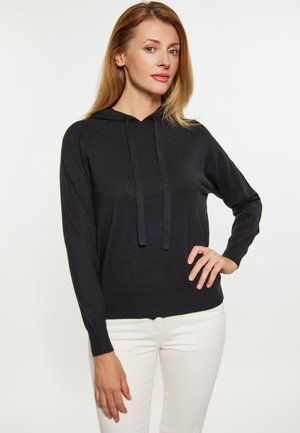 Frau mit hellbraunen Haaren, die einen schwarzen Hoodie und weiße Hose trägt, steht mit einer Hand in der Hüfte vor weißem Hintergrund.