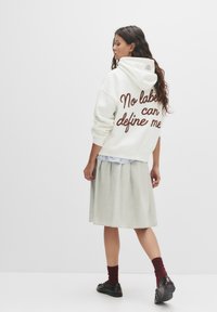 Felpa bianca con testo marrone "Nessuna etichetta può definirmi", abbinata a una gonna grigia chiara e scarpe nere, con una vestibilità rilassata e un grande cappuccio.