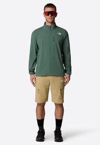 Vihreä kevyt takki, jossa on korkea kaulus ja vetoketjulliset taskut, yhdistettynä vaaleanruskeisiin shortseihin, joissa on sivuvetoketjut, sekä mustiin vaelluskengiin.