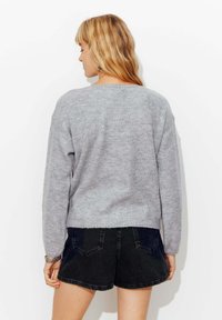 Femme blonde aux cheveux ondulés portant un pull en tricot gris clair et un short en jean noir, se tenant face à un mur blanc.