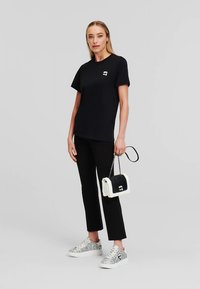 Mulher vestindo uma t-shirt preta e calças, segurando uma bolsa preta e branca e usando ténis prateados com brilho, de pé contra um fundo simples.