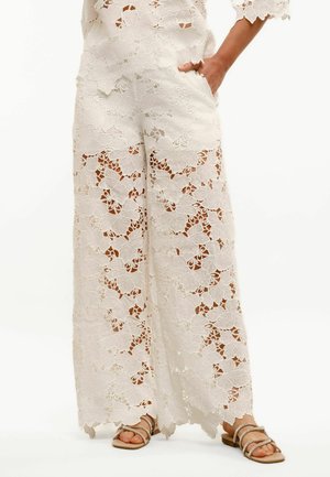 Białe spodnie o szerokich nogawkach z kwiatowym wzorem koronki, noszone z sandałami na paski i otwartym palcem, dłoń swobodnie w kieszeni.