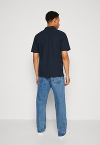 Camisa de botones de manga corta en color navy con un tejido texturizado, combinada con jeans rectos de color azul claro y zapatillas blancas.