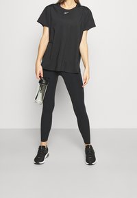 Musta lyhythihainen urheilupaita, jossa pieni logo, yhdistettynä mustiin leggingsseihin. Henkilö pitää kädessään kirkasta vesipulloa.