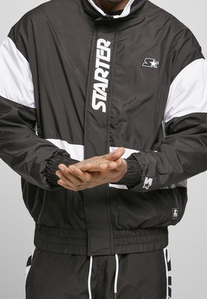 Homme portant une veste coupe-vent Starter noire et blanche avec logo sur la poitrine et la manche, les mains jointes devant lui.