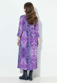 Femme aux longs cheveux ondulés portant une robe midi à manches longues à motifs violets et des bottes noires à lacets, se tenant de dos.