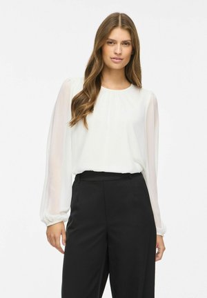 Jeune femme aux longs cheveux bruns ondulés, vêtue d'un chemisier blanc à manches longues et d'un pantalon noir taille haute, se tenant devant un fond blanc uni.
