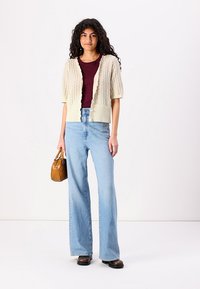 Knit cream cardigan with ribbed hem, short sleeves, and button closure, paired with high-waisted light blue wide-leg jeans and brown ankle boots.