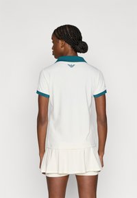 Λευκό φόρεμα πόλο με φαρδιά τελείωμα, με τιρκουάζ γιακά και μανσέτες. Το λογότυπο στο πίσω μέρος προσθέτει λεπτομέρεια. Ομαλή υφή υφάσματος.