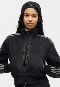 Νεαρή γυναίκα φοράει μαύρο τζιν μπουφάν Adidas με λευκές ρίγες και μαύρο καπέλο φορά με το ανάποδο, κοιτάει μπροστά με τα χέρια στις τσέπες.