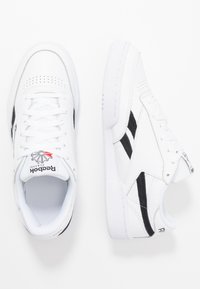 Biele kožené tenisky s čiernymi akcentmi, perforovaným dizajnom prstov, textúrovanými stranami a gumovou podrážkou. Obsahujú logo Reebok na jazyku.