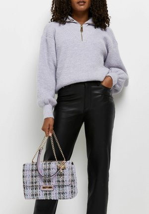 Kvinde iført en lys lilla lynlåstrøje og sorte læderbukser, der holder en lilla og sort ternet håndtaske med gyldne kæderemme.