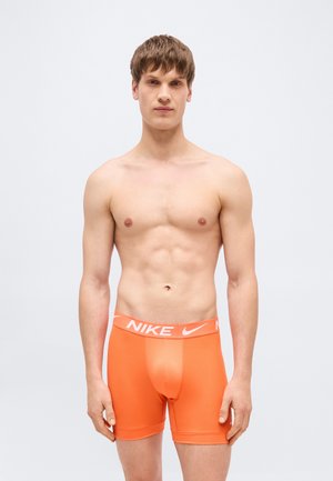 Jonge man staand, draagt feloranje Nike boxershorts, tegen een effen lichtgrijze achtergrond, met het gezicht naar voren en ontspannen armen.