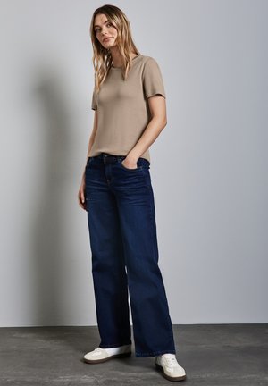 Beige short-sleeve t-shirt paired with dark blue wide-leg jeans and white sneakers, on a model against a gray background.