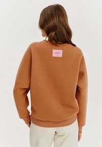 Hnědá oversized mikina s kulatým výstřihem, žebrovanými manžetami a růžovou nášivkou na zádech. Měkká textura a uvolněný střih.