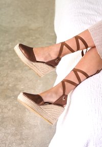 Alpargatas cuña de cuero marrón con suela de cuerda de yute trenzada, diseño con tiras cruzadas y detalle circular de metal en el talón.