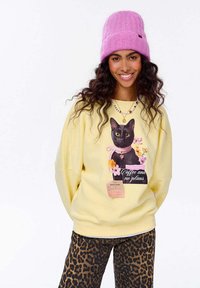 Žlutá mikina s grafikou černé kočky a květinovými akcenty a textem "Káva a žádné plány". V kombinaci s kalhotami s leopardím vzorem a růžovou pletenou čepicí.