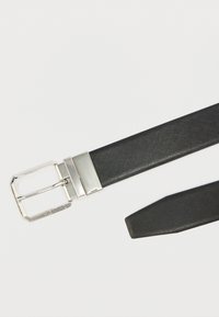 Fekete, texturált öv ezüst téglalap alakú csattal. A csaton márkajelzés található, és sima felületű. Az öv vége felé keskenyedik.