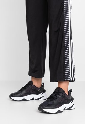 Person iført sorte og hvide chunky Nike sneakers med sorte bukser med hvide stribede sidepaneler.