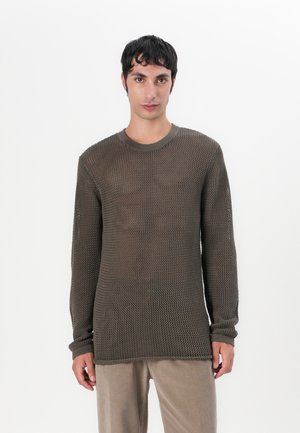 Pulover lung, tricotat, de culoare maro, cu un design texturat, cu o țesătură deschisă. Prezintă un guler rotund și manșete îndoite, purtat cu pantaloni bej.