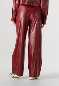 Persoon die wijde, hooggetailleerde bordeauxrode leren broek en een bijpassend jasje draagt, met witte en grijze sneakers, staande op een witte vloer.