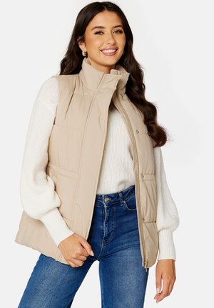 Beige gewatteerde mouwloze vest met een opstaande kraag en drukknoopsluitingen. Beschikt over voorzakken, een gladde textuur en lichte isolatie. Draag het over een witte trui.