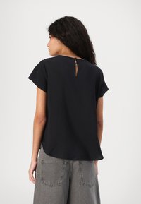 Blusa nera a maniche corte con dettagli sulla schiena a chiave, caratterizzata da una vestibilità ampia e una texture morbida. Abbinata a pantaloni grigi a gamba larga.