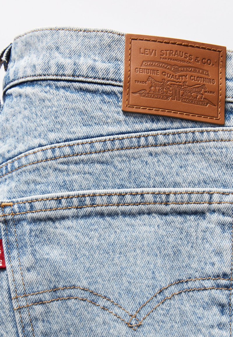 Jeans en denim bleu clair avec une partie en cuir tan portant des graphismes en relief, des coutures dorées et une étiquette rouge signature sur la poche arrière.