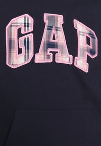 Námořnická mikina s nápisem "GAP" v velkých písmenech stylu plaket. Písmena mají růžový okraj a kostkovanou texturu v odstínech modré a růžové.