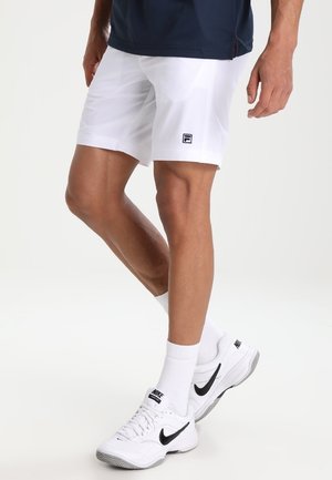 Mężczyzna w białych szortach Fila, białych butach Nike z czarnym logo Swoosh oraz białych skarpetach, stojący na jednolitym białym tle.