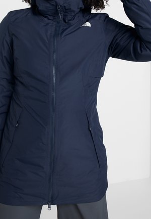 Femme portant une veste North Face bleu marine à fermeture éclair avec poches latérales, vue partielle des épaules aux hanches sur un fond uni.