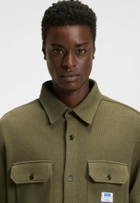 Olive green ribbed shirt with a collar, featuring two front pockets, black buttons, and a logo label on the left side.