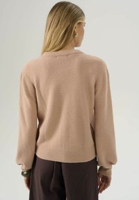 Donna con lunghi capelli biondi, che indossa un maglione beige con texture e pantaloni marroni scuri, di spalle contro uno sfondo semplice e chiaro.