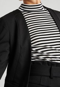 Veste noire portée sur un col roulé rayé noir et blanc. Gros plan montrant les textures du tissu et les motifs de couleurs contrastantes.