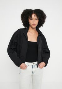 Mustast lõhestatud bomberjakk kõrge kraega, pikkade varrukatega ja siledast, läikivast materjalist. Kandmise all must top ja heledad teksad.