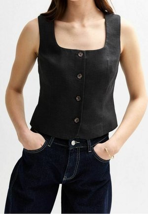 Žena v bezrukávovém černém topu s knoflíky a čtvercovým výstřihem, tmavě modré džíny, ruce v kapsách, na jednobarevném pozadí.