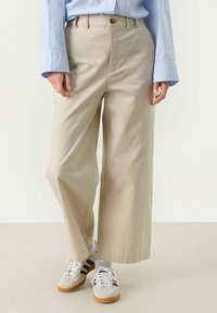 Beige wide-leg trousers with a high waist, front button detail, and soft texture, paired with light-colored sneakers with black stripes.