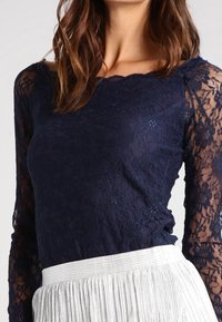 Haut en dentelle bleu marine à manches longues avec un décolleté festonné, associé à une jupe plissée de couleur claire. Motif floral texturé sur les manches.