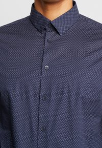 Marinblå knapp-up skjorta med ett litet vitt geometriskt mönster, med klassisk krage och slät textur, tillverkad av ett lättviktigt tyg.