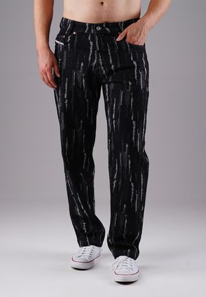 Mand iført sorte jeans med grå lodrette striber og hvide sneakers, står med den ene hånd i lommen mod en grå baggrund.