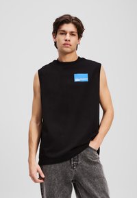 Črna brezrokavna majica z okroglim izrezom in modrim pravokotnim logotipom na levi strani, v kombinaciji s sivimi jeans hlačami.