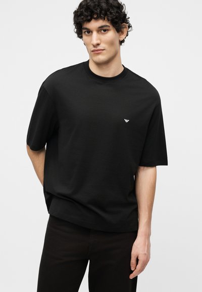 Νέος άνδρας με σγουρά μαλλιά, φορώντας μαύρο κοντομάνικο μπλουζάκι με μικρό λευκό λογότυπο στο αριστερό στήθος, στέκεται μπροστά σε απλό φόντο.