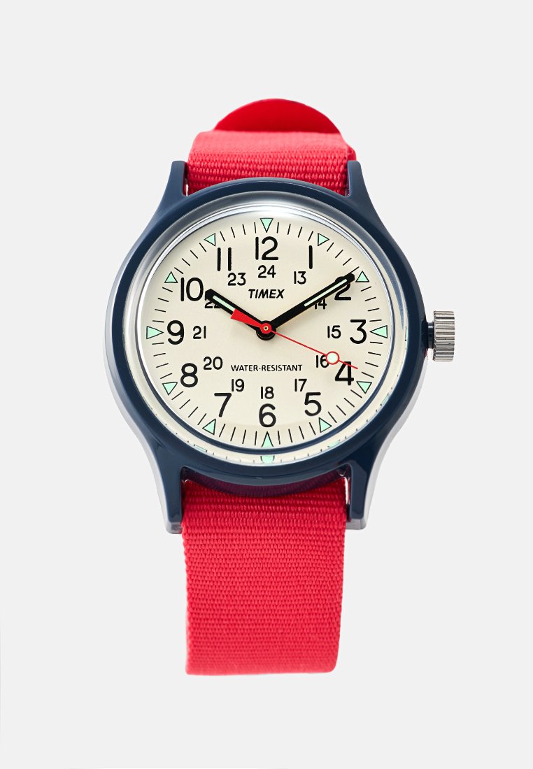 Reloj Timex con caja de plástico redonda en color azul marino, esfera blanca, agujas negras, correa de nailon roja y marcadores numéricos. Se señala la característica de resistencia al agua.