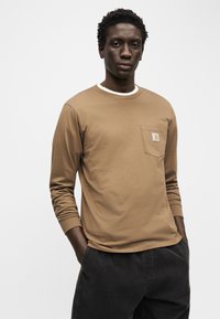 Νέος άνδρας με τα χέρια στις τσέπες, φορώντας καφέ μακρυμάνικο πουκάμισο με λογότυπο σε τσέπη στο στήθος και σκούρο παντελόνι, μπροστά από απλό φόντο.