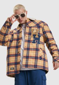 Tan plaid shirt jacket with blue and red accents, featuring a soft shearling collar and embroidered letters on the chest.
