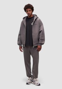 Felpa grigia con zip e due tasche frontali, abbinata a una t-shirt nera e a pantaloni grigi coordinati. Dettagli delle sneakers con rete e accenti audaci.