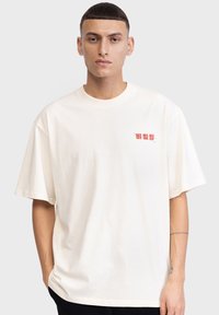 T-shirt en coton crème avec une coupe décontractée. Présente un petit texte graphique rouge en haut à gauche. Manches courtes et col rond.