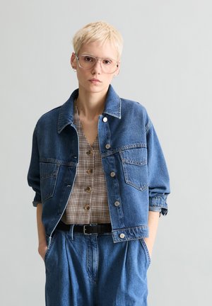 Osoba o krótkich blond włosach, ubrana w oversize'ową kurtkę jeansową, koszulę w kratę na guziki, spodnie jeansowe, okulary i czarny pasek, stojąca na tle szarego tła.