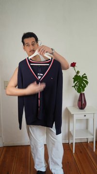 Čovjek drži tamnoplavi kardigan s crvenim i bijelim obrubom na vješalici, stojeći pored bijelog stola s crvenom vazom i jednom crvenom ružom.