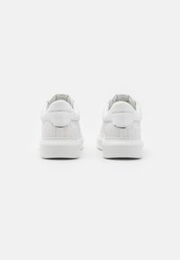 Hvite sneakers med en teksturert patentlærfinish, rund tå, polstret krave, og subtil logo-detail på baksiden. Tykk gummisåle.